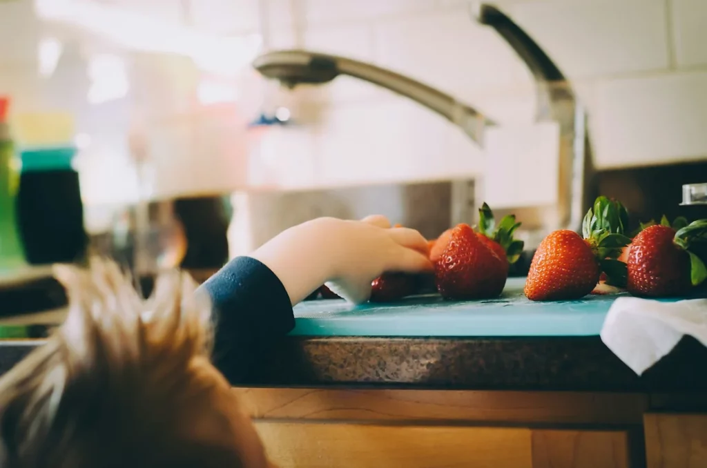 A child reaches up to grab a strawberry off a cutting board on the kitchen counter.